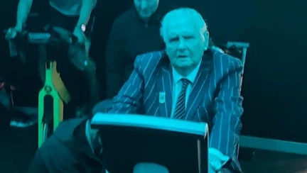 Norman is sat on an exercise bike wearing a striped blazer jacket and tie