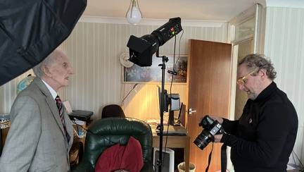Peter stands for a photograph in his living room, as Richard checks his camera. Lighting equipment and a black backdrop are seen in the background.