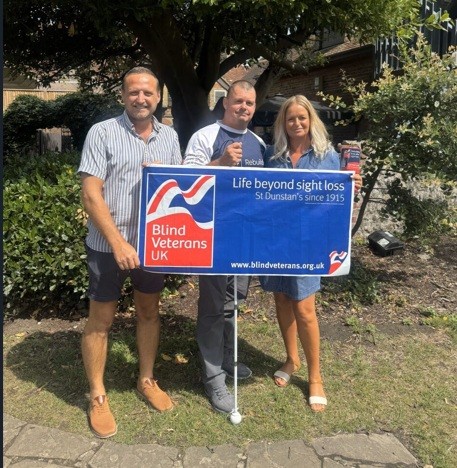 Stu with his Blind Veterans UK t-shirt and holding his white cane is stood between Heidi and Niall from The Bull and the three of them are holding a Blind Veterans UK banner
