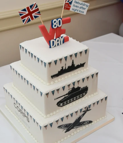 A three tiered celebration cake decorated with red, white and blue bunting and silhouettes of military vehicles. The top features a fondant VE Day 80, a Blind Veterans UK flag and a Union Jack flag.