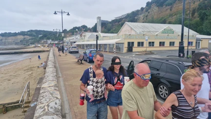 A lady has a blindfolded walker holding on to each of her arms and behind them is a man with a baby in a carrier who is guiding a blindfolded lady in a Blind Veterans UK T-shirt