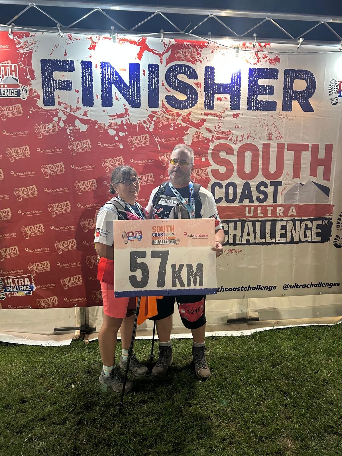 Andy and Angie holding a banner saying 57km with a large backdrop saying 'Finisher'