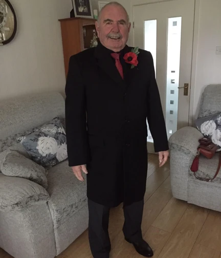 Joe is stood inside a living room wearing a smart black coat, red tie and large poppy