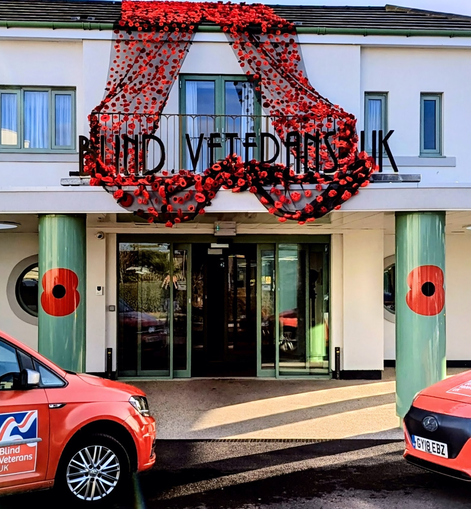 A display of knitted poppies draped over the entrance of Blind Veterans UK's Rustington Centre, with poppy decorations on the columns and two Blind Veterans UK vehicles parked outside.