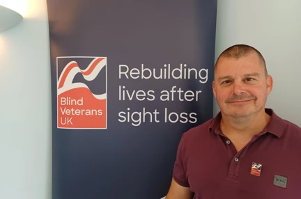 Stuart looking straight at the camera and smiling stood next to a Blind Veterans UK banner