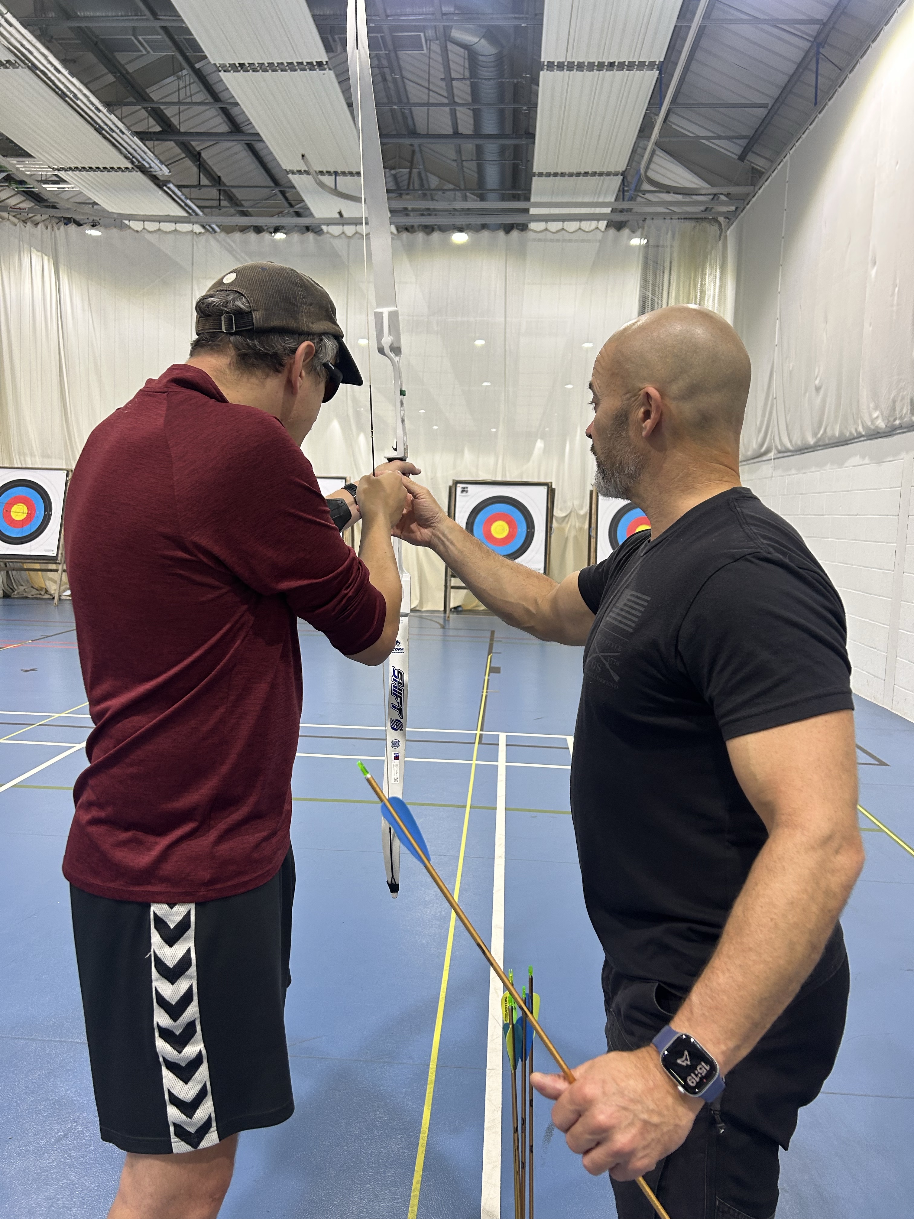 A blind veteran holds a bow and points it towards a target. A staff member shows him how.