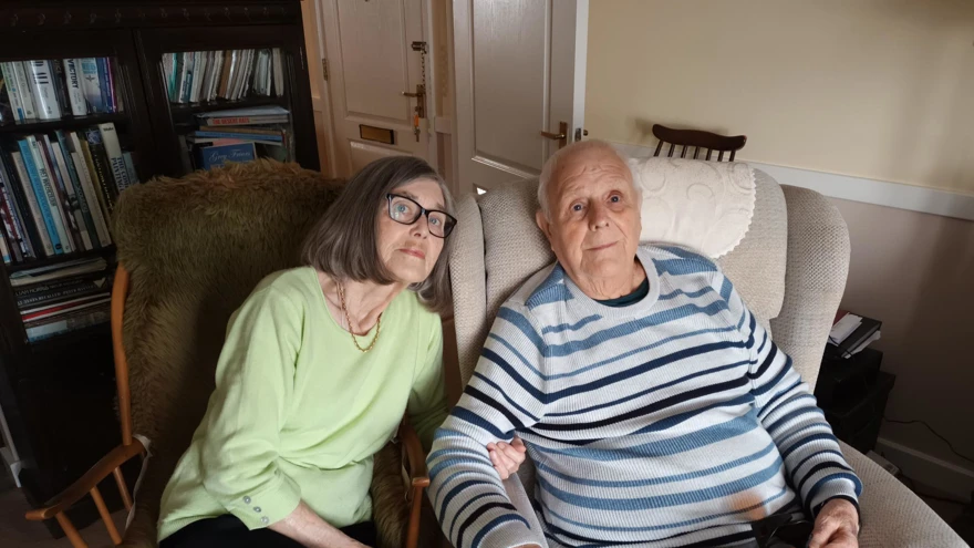 A photo of blind veteran Dudley, right, and his wife Yvonne, left sitting on chairs while linking arms
