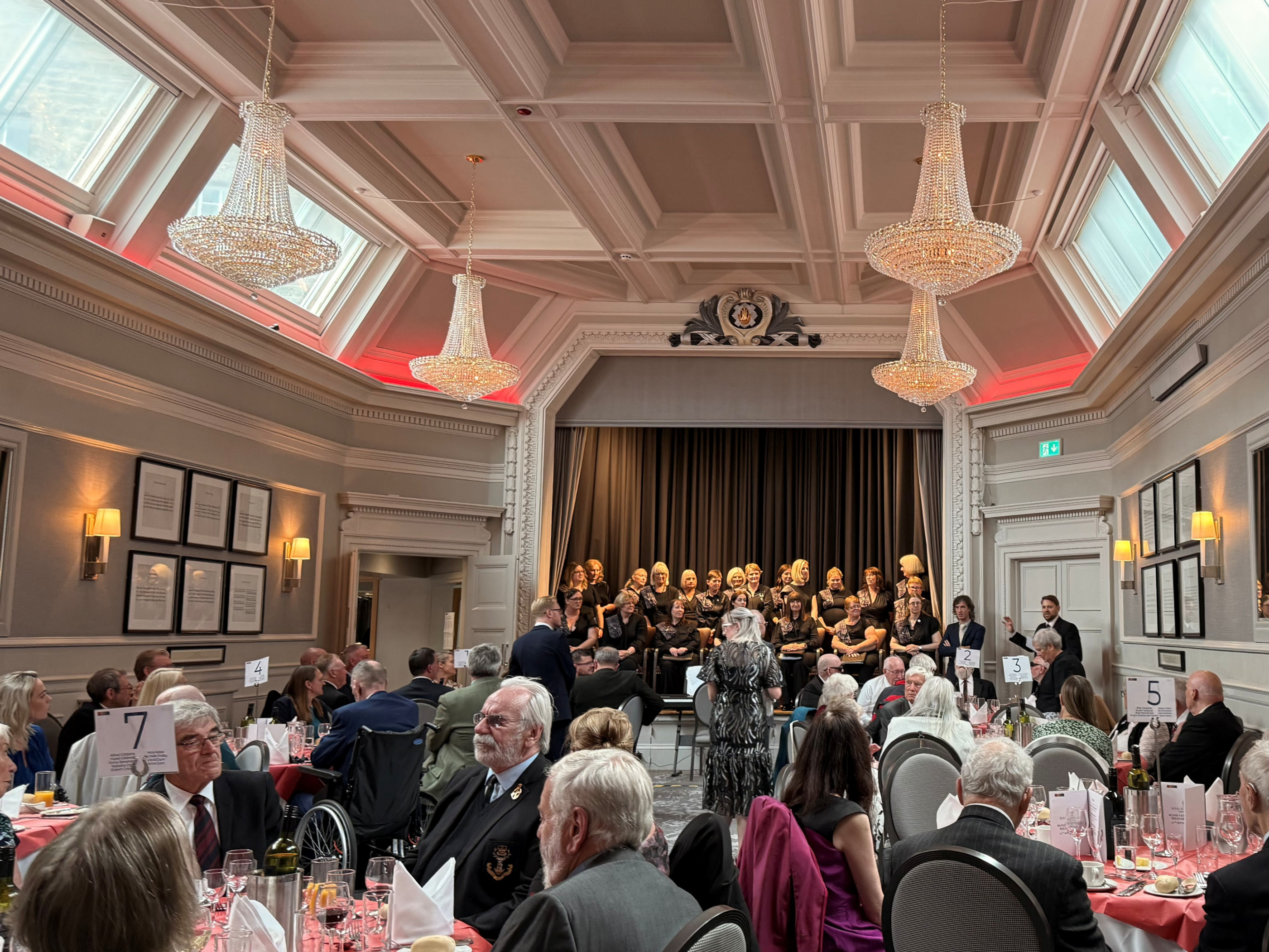 A wide shot of an event taking place. People are seated at round tables and a choir is singing on stage.