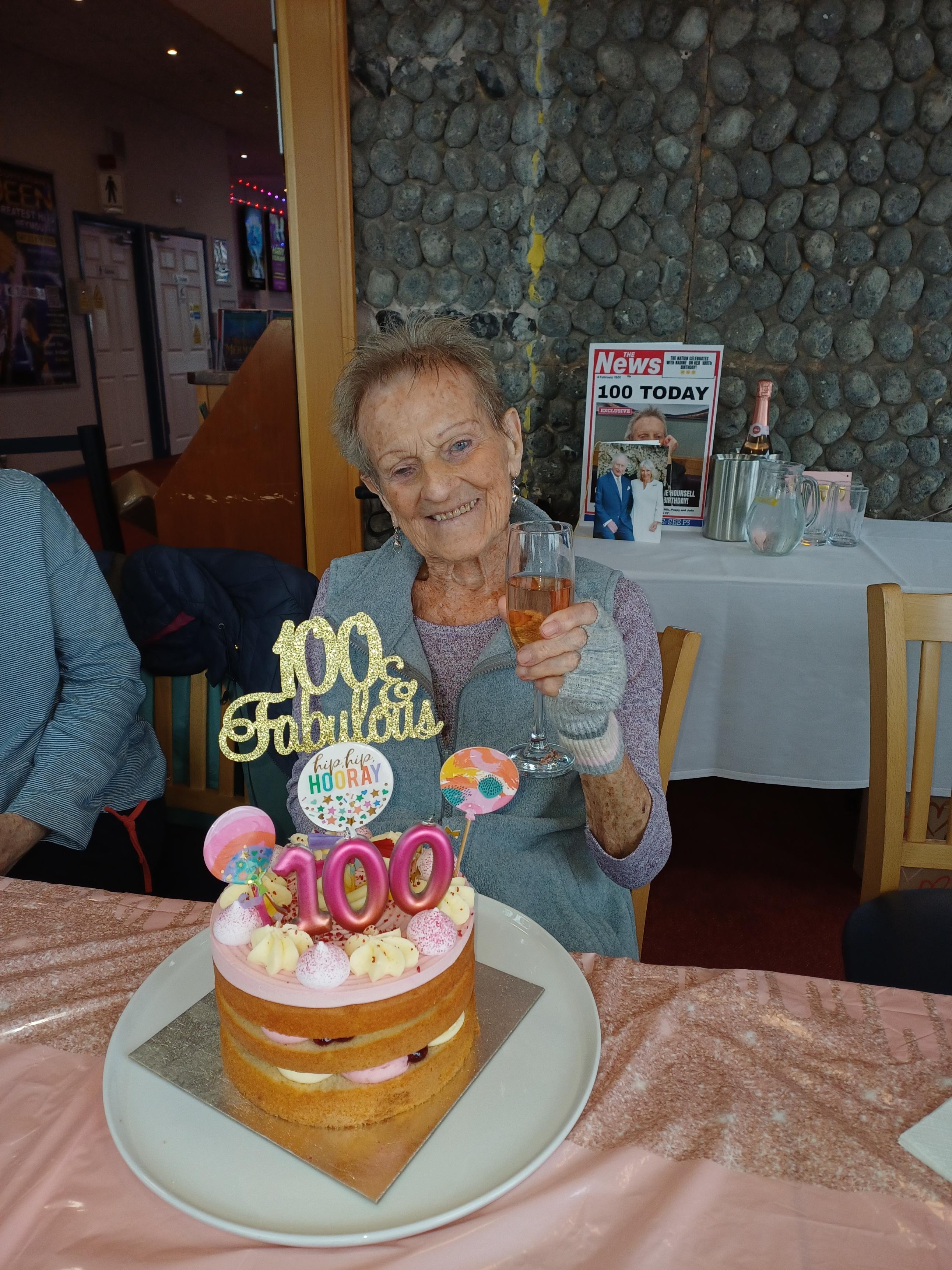Image shows blind veteran Nadine celebrating her 100th birthday, seated before her cake and raising a champagne glass