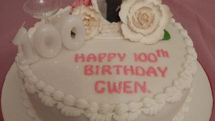 A cake with white frosting, 100th birthday candles, floral detail and a picture of blind veteran Gwen placed on top