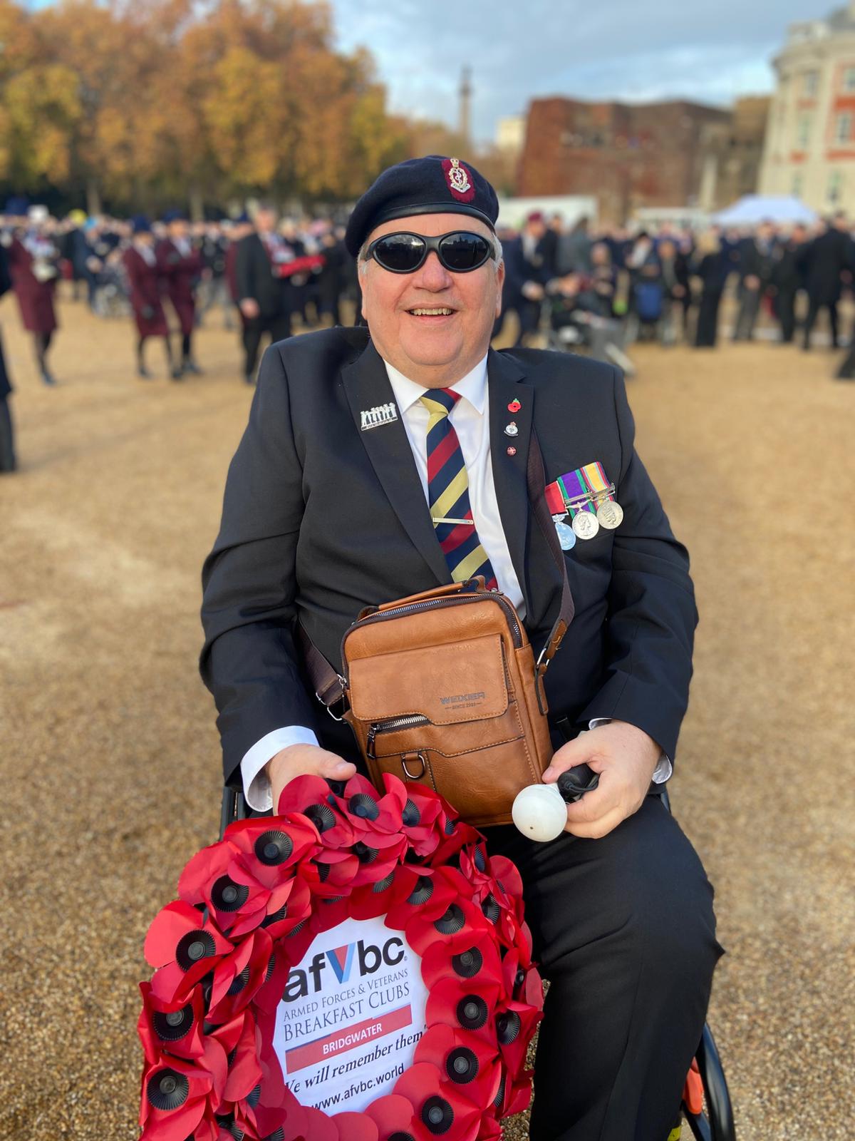 John sits in a wheelchair and looks into camera, holding an Armed Forces Breakfast Club wreath