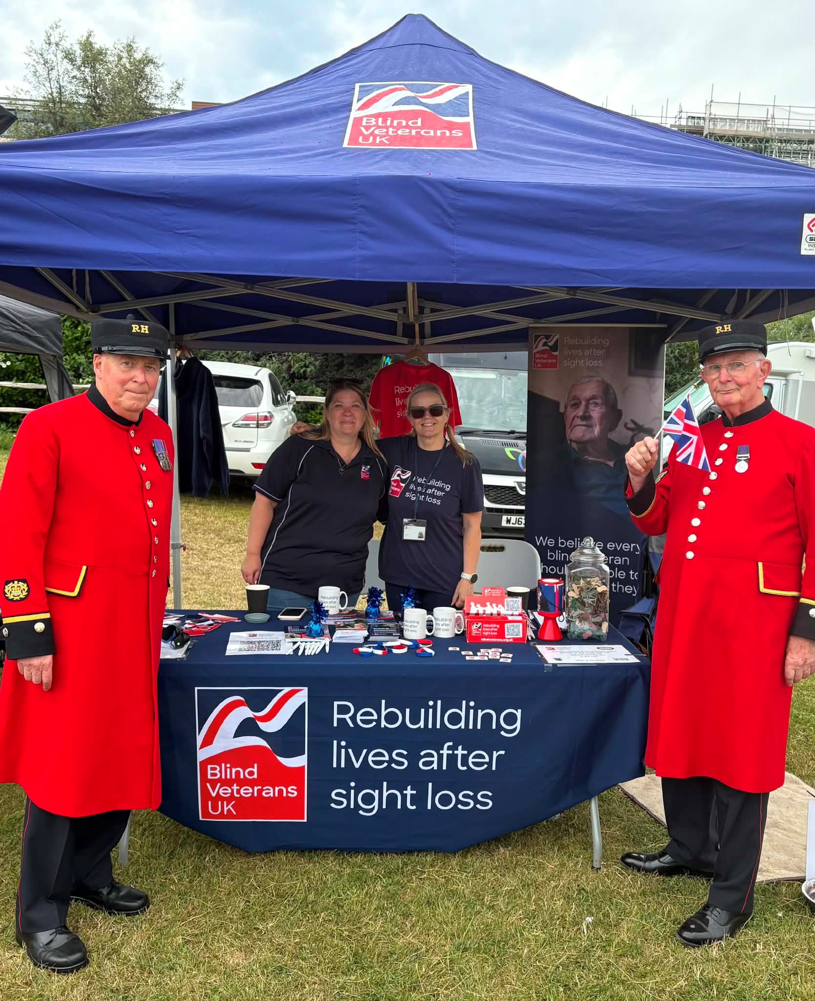 Two members of staff in their Blind Veterans UK tops under a branded Gazebo.  Two Chelsea Pensioners are stood either side of the Gazebo.