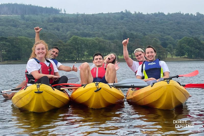 A group of event participants in two-person kayaks on the lake, with tree-covered hills in the background. The group are all smiling at the camera.  Some are holding their arms up while signalling a thumbs up.