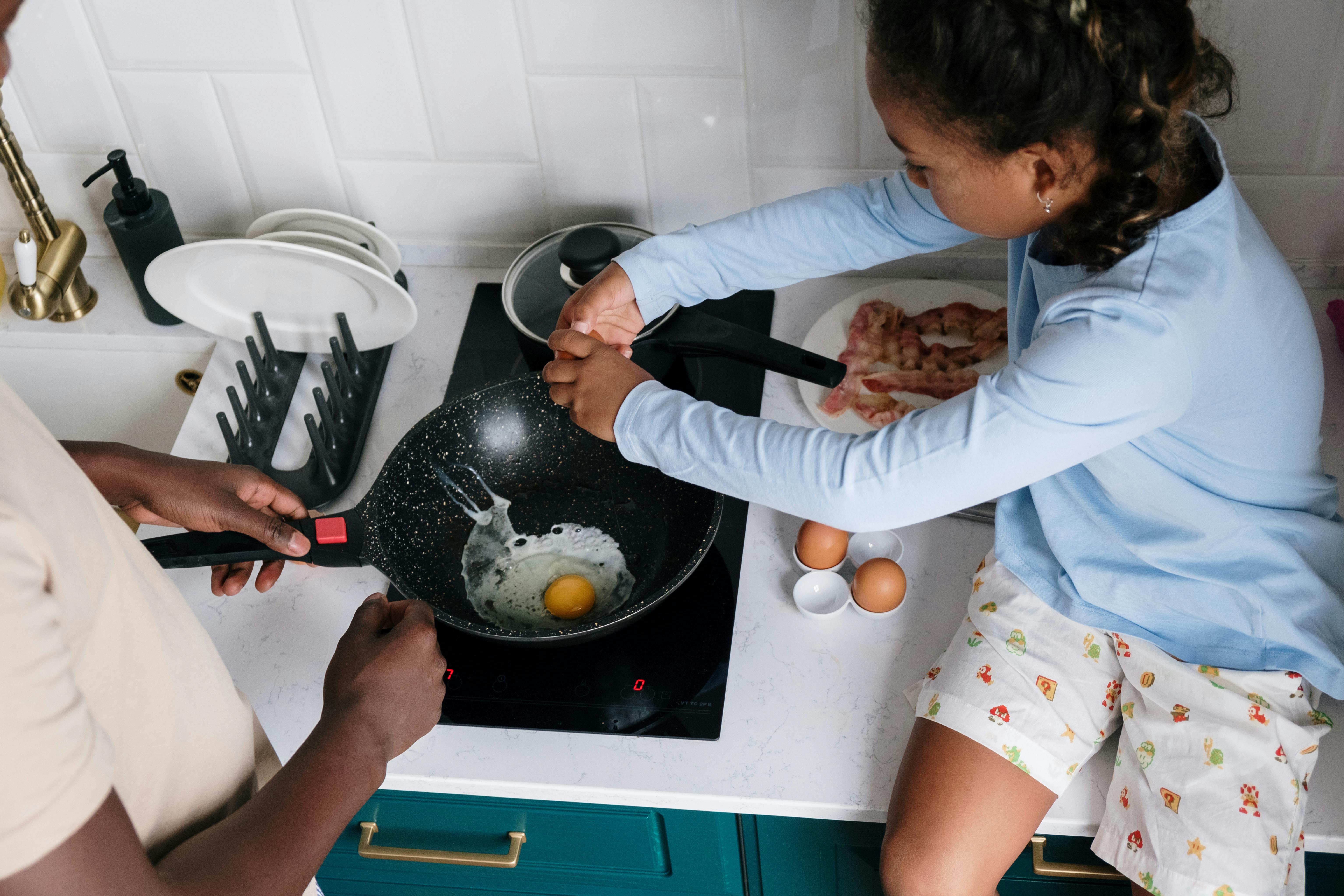 Two people cooking at a kitchen hob; one cracks an egg into a frying pan while the other holds the pan handle, with eggs, bread and kitchen items arranged on the countertop nearby.