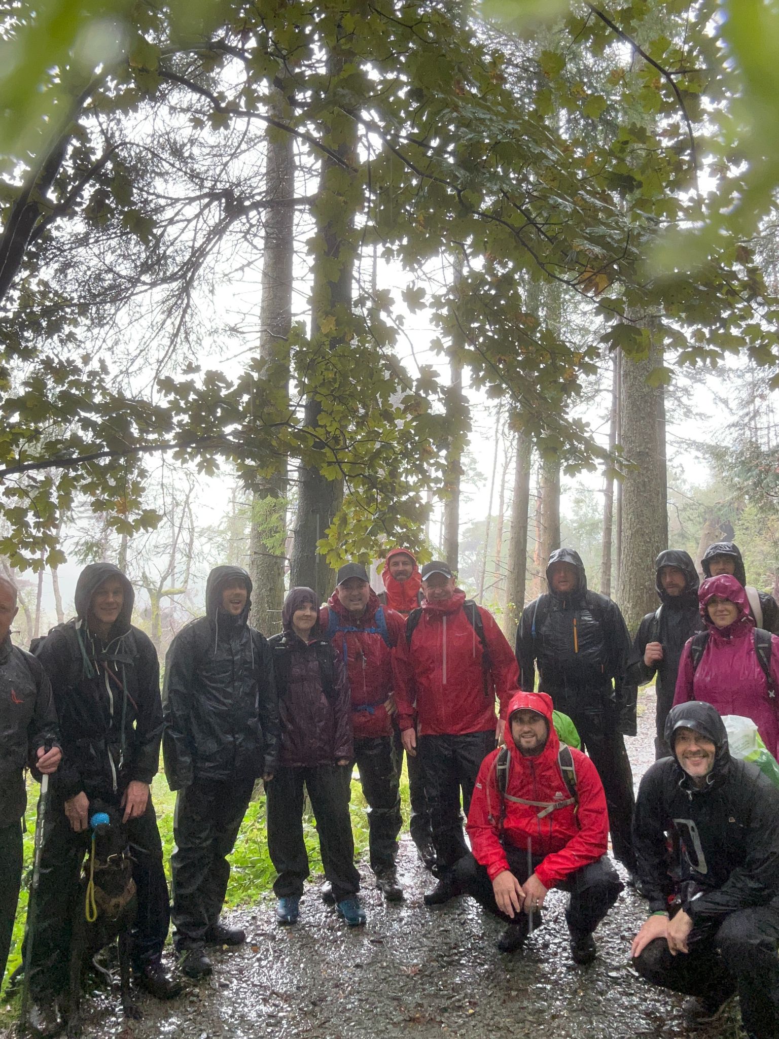 A group picture of the walkers paused under some trees. The ground is wet and they are soaked. Fully kitted out in waterproof coats and waterproof trousers.