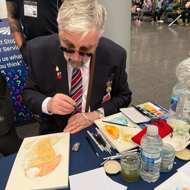 Bryan dressed smartly in jacket and Blind Veteran UK tie sat at a table painting with watercolours