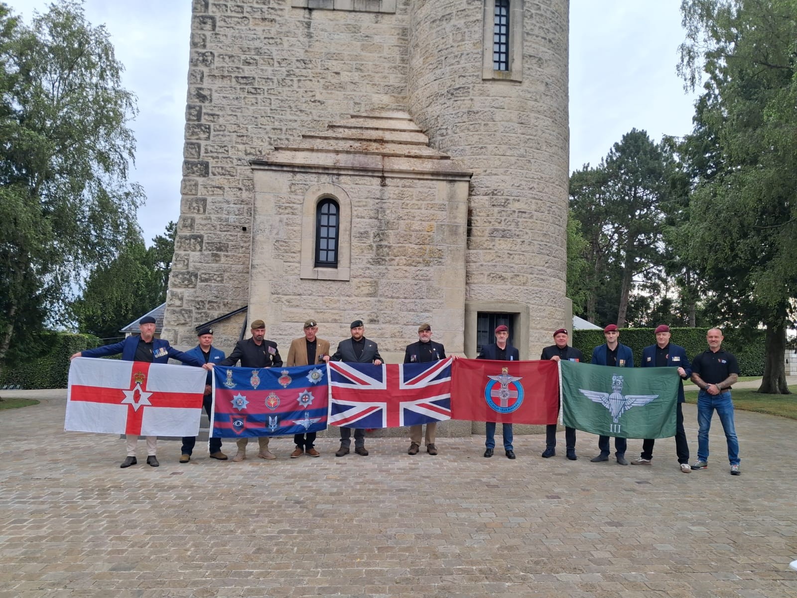 11 veterans stand next to each other, all wearing berets and holding regiment flags