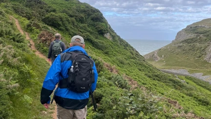 A picture of Phil from behind as he uses his white cane to navigate along a thin pathway which is going uphill along the coastline