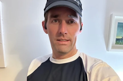 Chris is stood inside his house wearing his Blind Veterans UK running top and a baseball cap