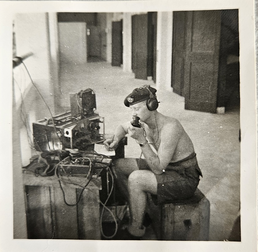 Arthur sits in front of radio equipment with his beret and headphones on, he speaks into a mouthpiece