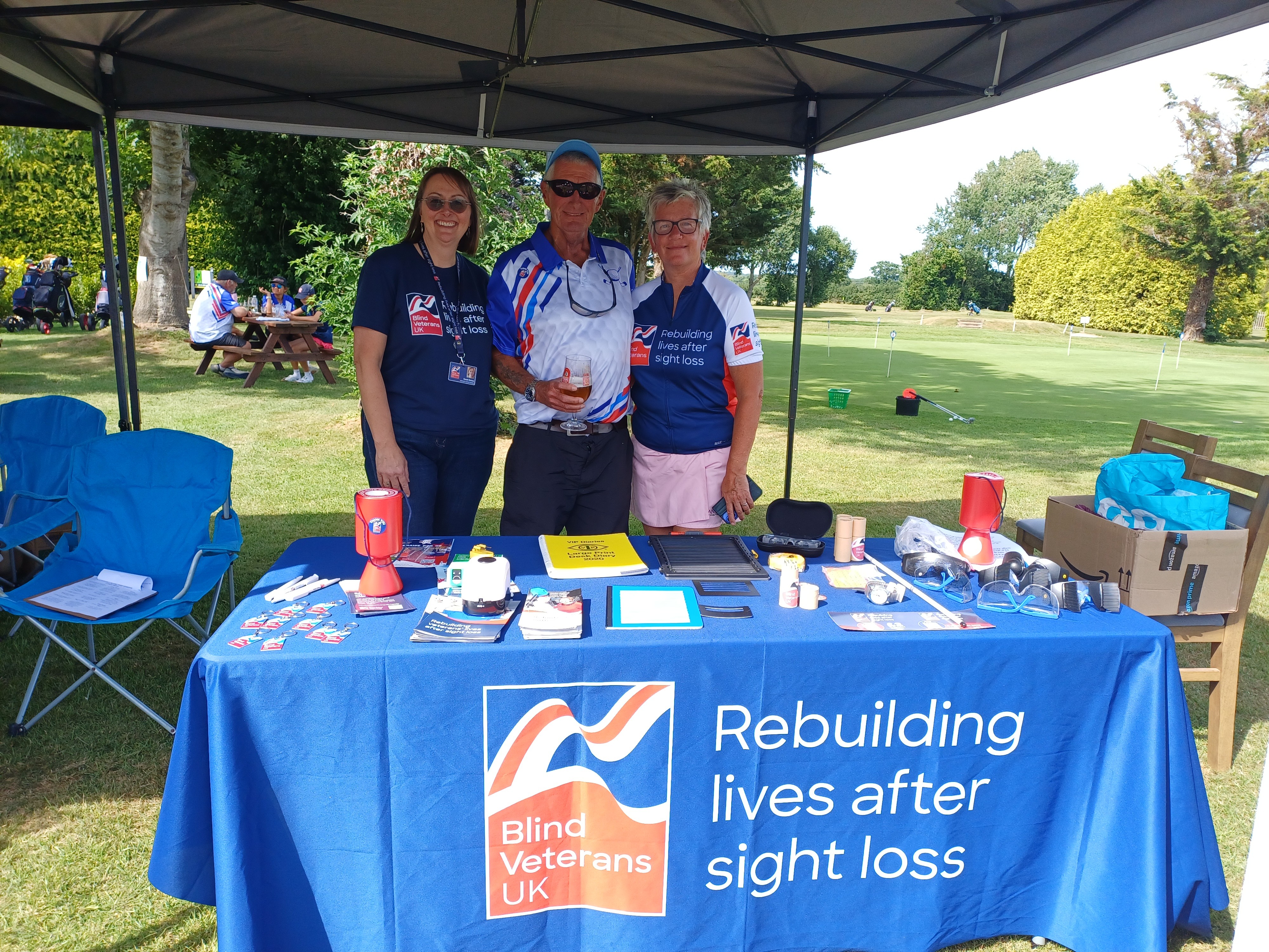 Tony, Kozy and Sarah stood at the Blind Veterans UK information table which has lots of leaflets and the simulation spectacles 