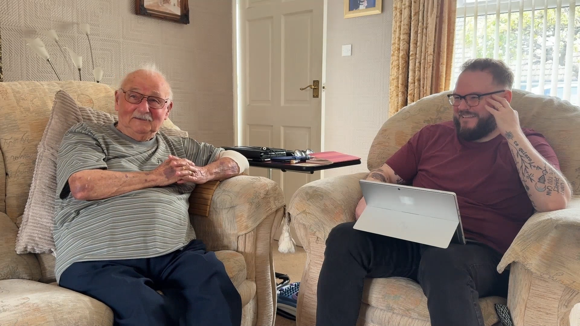 Ken and David sit in armchairs during a home visit, with David holding a tablet as they talk.