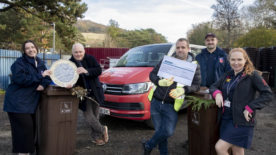 A picture of people leaning against two brown wheelie bins. One group are holding a cheque and the other a large £1 coin.