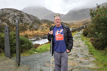 Phil on a cobble stone path with mountains behind him. He has yellow tinted glasses on his head and is holding his long cane. He is wearing a Blind Veterans UK T-shirt, grey walking trousers and a black jacket.