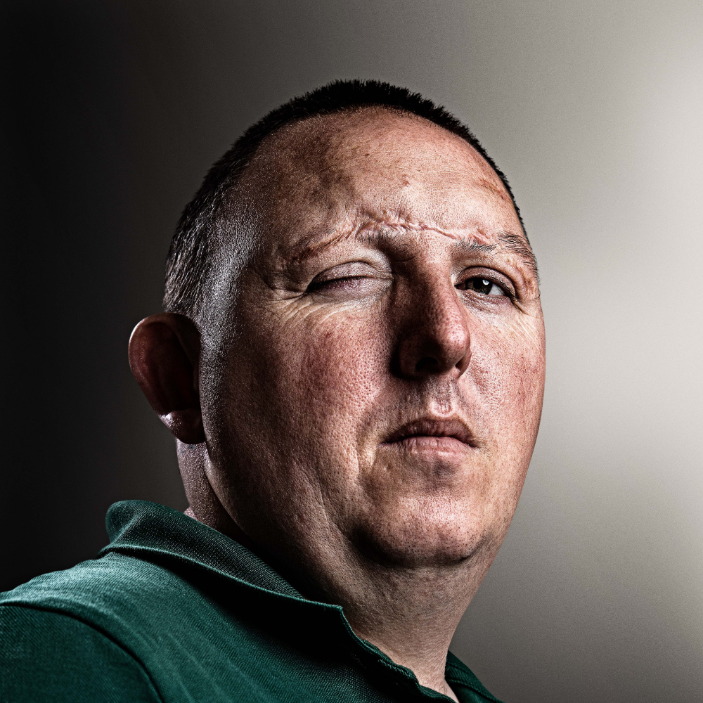 Portrait of blind veteran Chris in a green shirt against a neutral background.