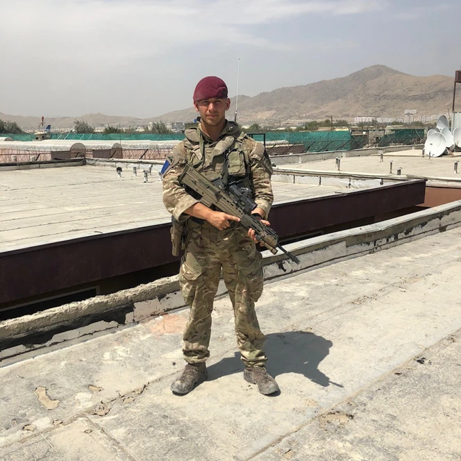 Jacob dressed in military camouflage and a maroon beret, stands on a rooftop holding a rifle. The background shows a mountainous landscape in Kabul.