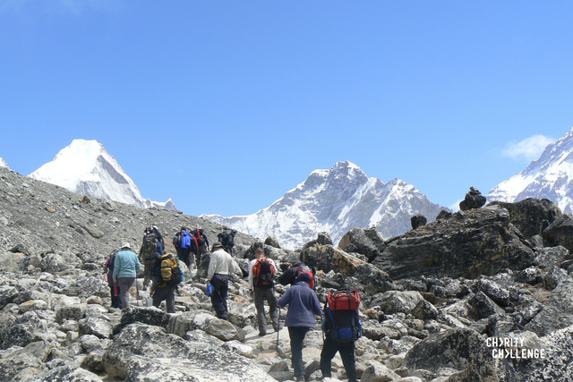 Group of people walking away from the camera across rocky snow-covered landscape, heading towards towering, icy mountains that fill the background of the image.