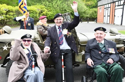 Blind veterans John, Harry and Ted smiling and waving in front of a historic military vehicle at the Petwood Hotel for VE Day 80.