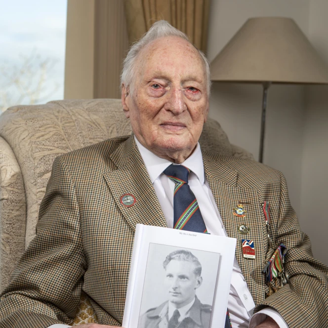Blind veteran Derrick seated in a chair holding a book with a photo of himself in service on the cover