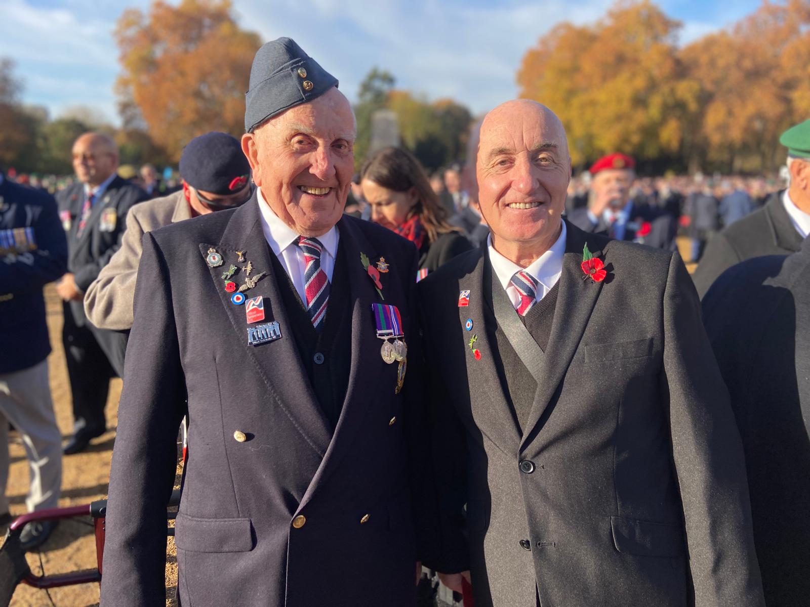 Blind veteran Michael and Eddie stand together at the Cenotaph, wearing formal suits, striped ties, poppies and military medals. Other attendees and autumn trees are visible behind them.