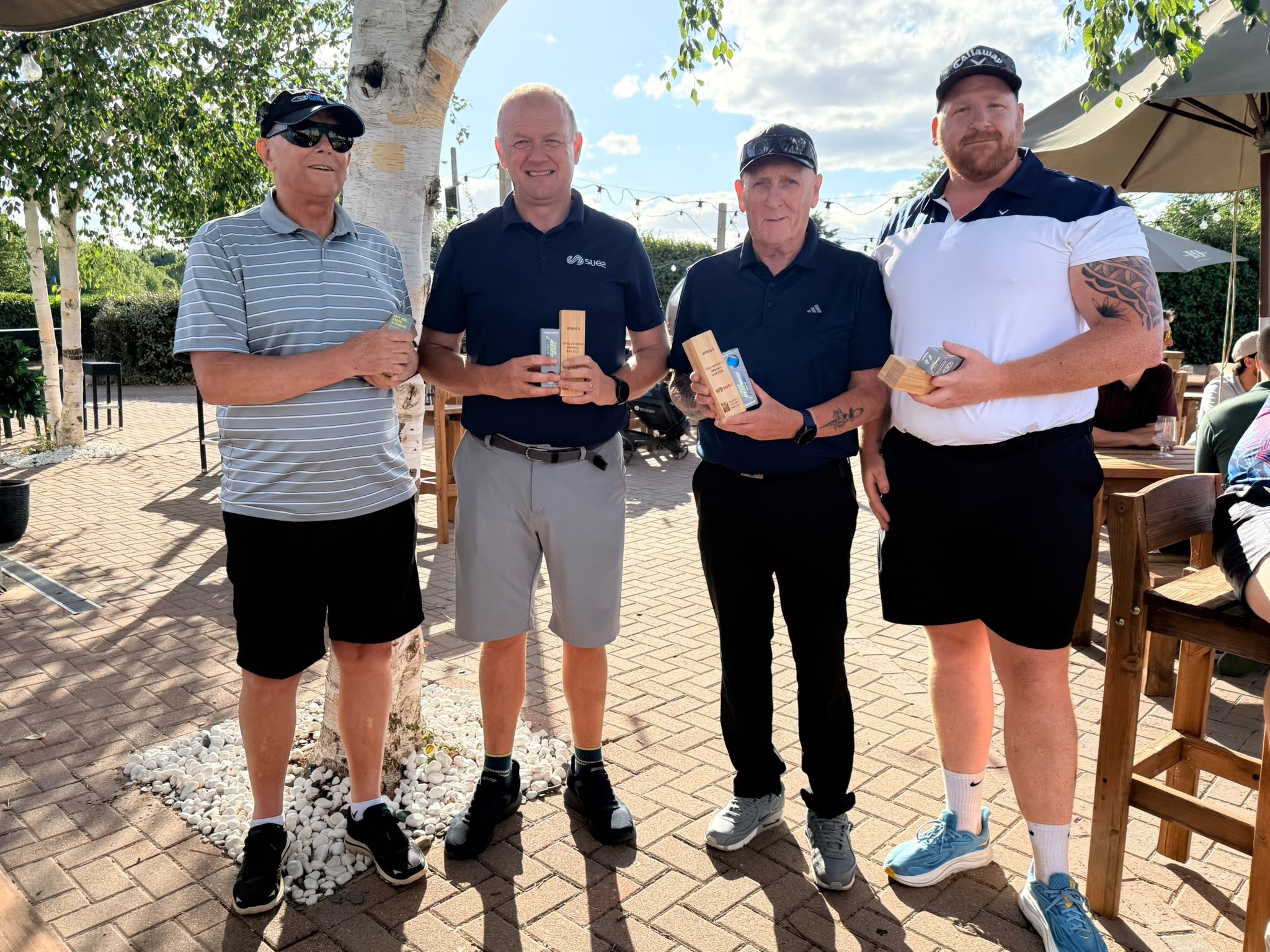 Four members of the winning golf team stand together outside the clubhouse holding their wooden trophies.