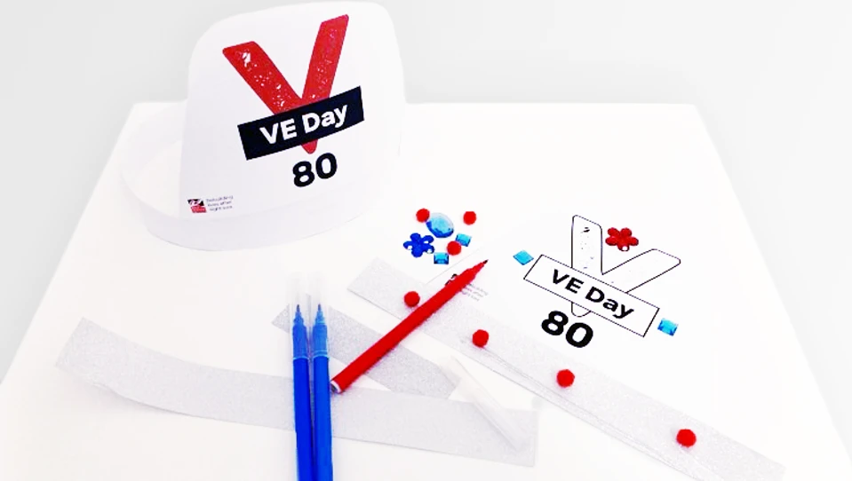 Assembled VE Day 80 party hat and one in the progress of being decorated with pom-poms, craft jewels and silver paper.