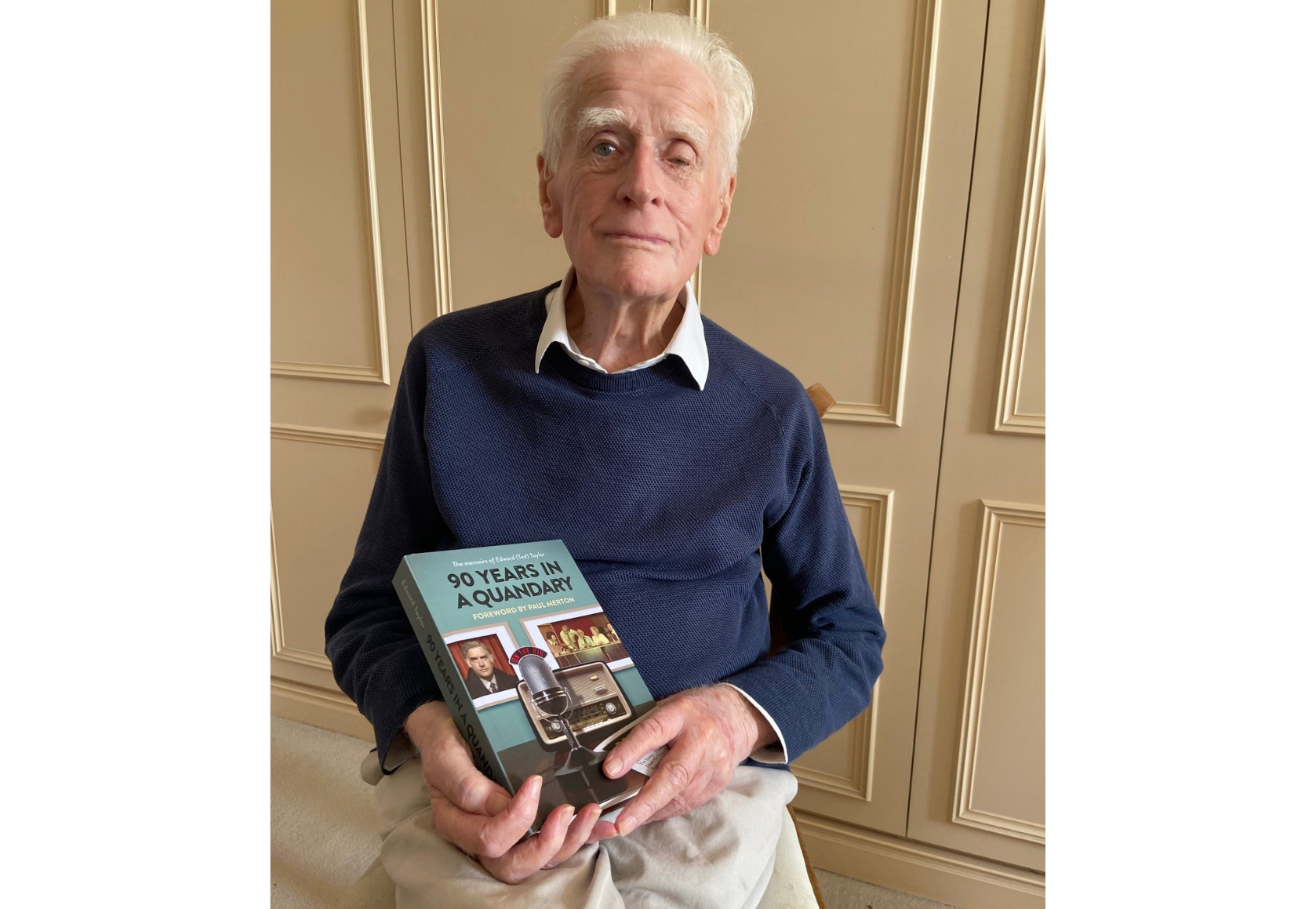 Ted sits on a chair and looks at the camera, holding the book in his hands