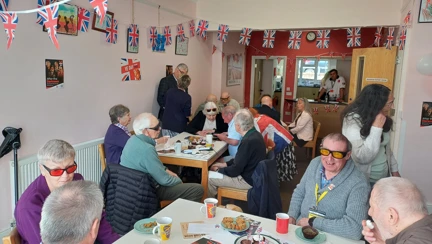 A group of blind veterans and supporters seated around tables decorated with red, white and blue bunting, enjoying a VE Day tea party.