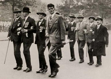 Black-and-white archive photograph of a group marching in formation. Several people wear medals, some use white canes and one person provides guided support near the front.