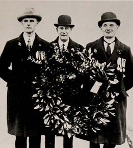 Black-and-white archive photograph of three people wearing dark coats, hats and military medals, holding two large wreaths made from leaves and flowers.