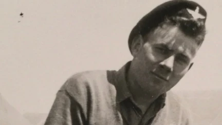 A black and white photograph of Danny during his military days. He is pictured in front of tents wearing a jumper and a military cap