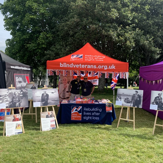 Two members of our community team standing underneath a Blind Veterans UK gazebo, and behind a stall decorated with merchandise and D-Day portraits