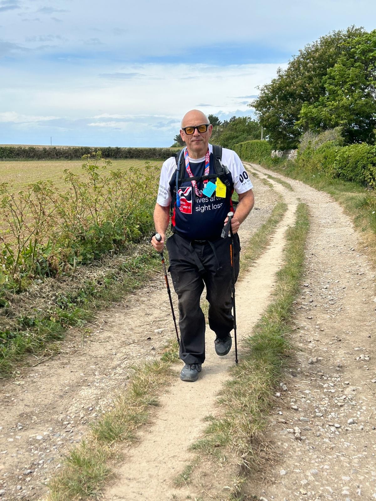 Jerry walking along a dirt track during. He is wearing yellow glasses and a Blind Veterans UK Tshirt and using walking poles