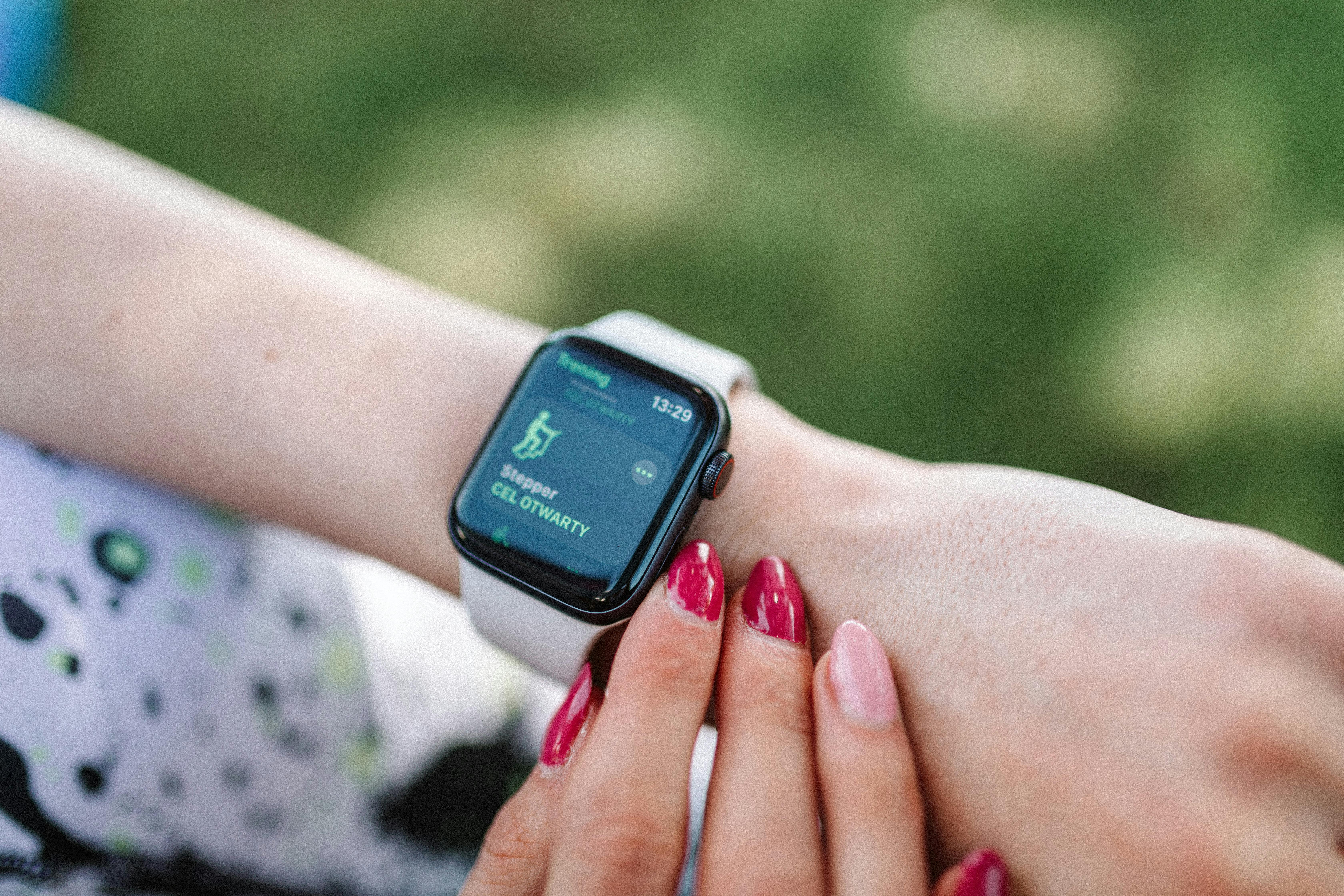 Close-up of a wrist wearing a smartwatch displaying step count, with another hand touching the screen; fingernails are painted and the background shows a blurred green outdoor setting.