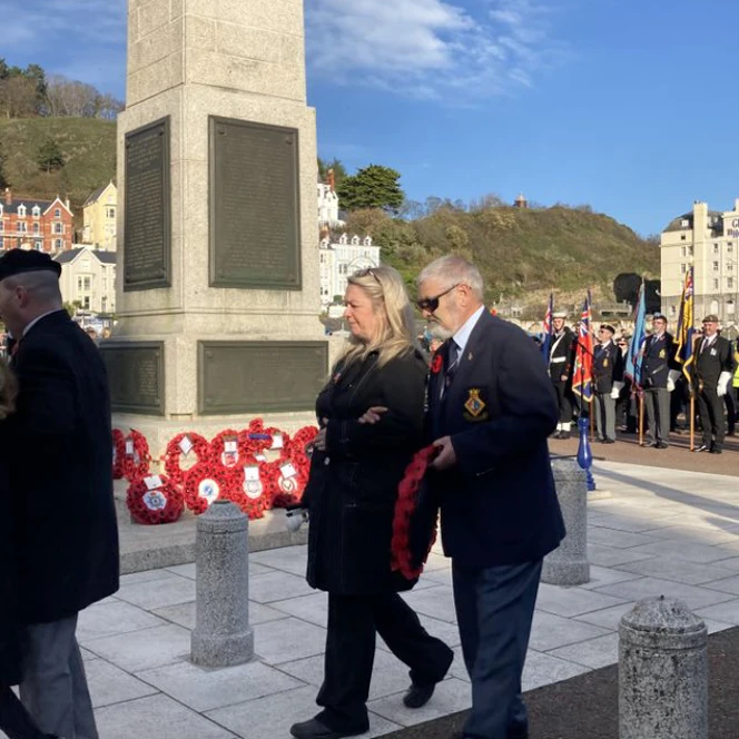 Bob wearing a suit and dark glasses is stood in front of the Llandudno War Memorial waiting to lay a wreath alongside those already placed