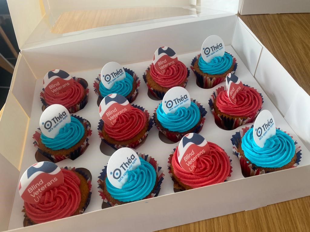 A box of cupcakes made by Thea Pharmaceuticals team members.  The cakes are each iced in blue or red butter cream, with Blind Veterans UK and Thea Pharmaceuticals logos on top in sugar paper.