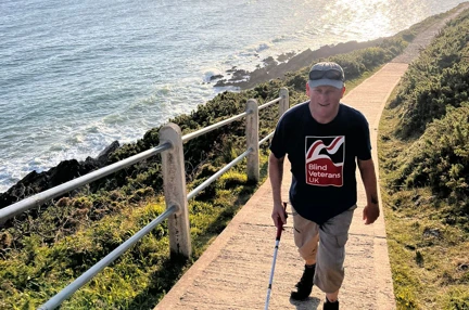 Phil walks along a thin concrete path along the coast with the sea to the side of him and the sun beginning to set behind