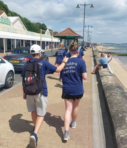 An image of the back of two walkers as they make their way along the seafront. A woman is acting as the guide and a man is holding on to her shoulder and walking slightly behind