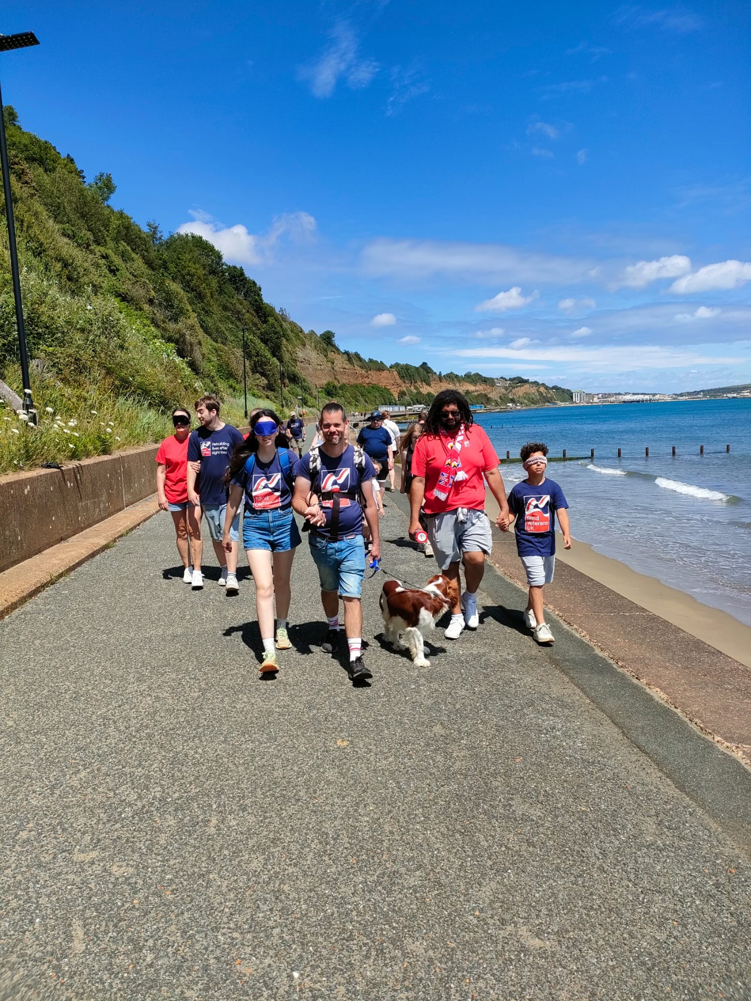 The group make their way along the promenade in the sun with the sea to one side of them 