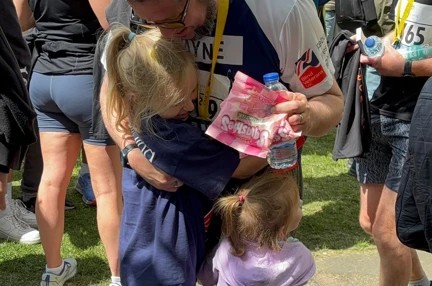 Wayne receiving a hug from his young daughters and a packet of sweets after finishing the race. He has his medal around his neck.